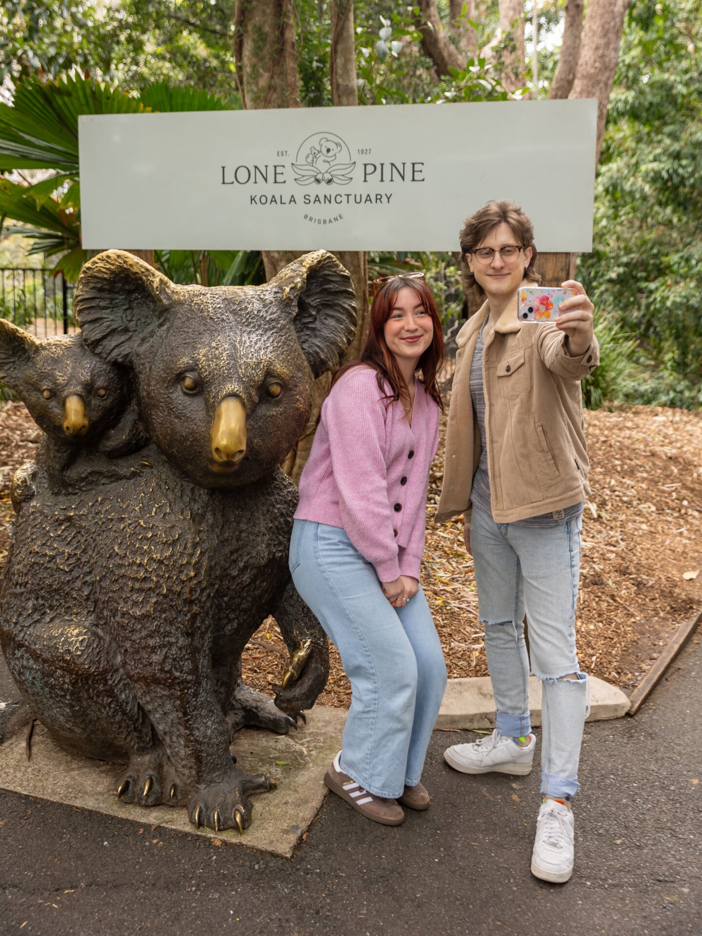 Two visitors taking a selfie beside the koala statue at the Lone Pine Koala Sanctuary entrance sign.