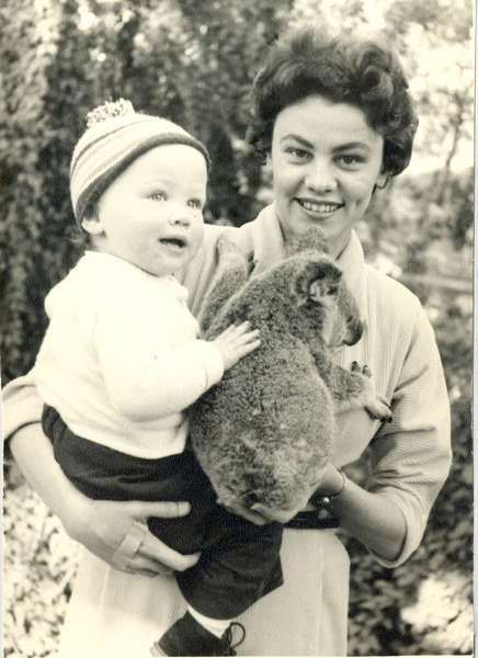 A smiling mother holding her baby in a knitted hat alongside a koala at Lone Pine, 1958